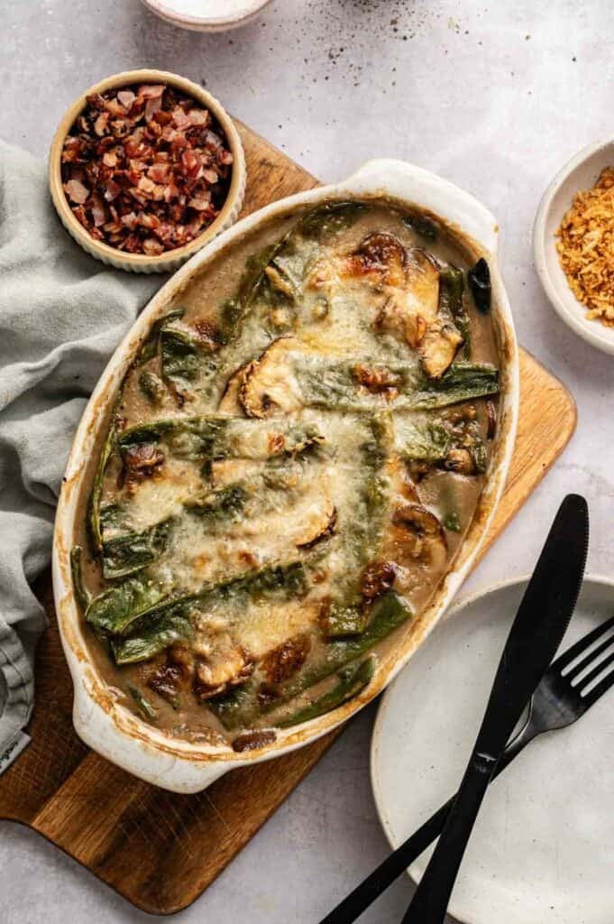 A baked Green Bean and Bacon Casserole topped with melted cheese, green beans, and sliced mushrooms, served in an oval dish on a wooden board with a knife, fork, and small bowls of toppings nearby.