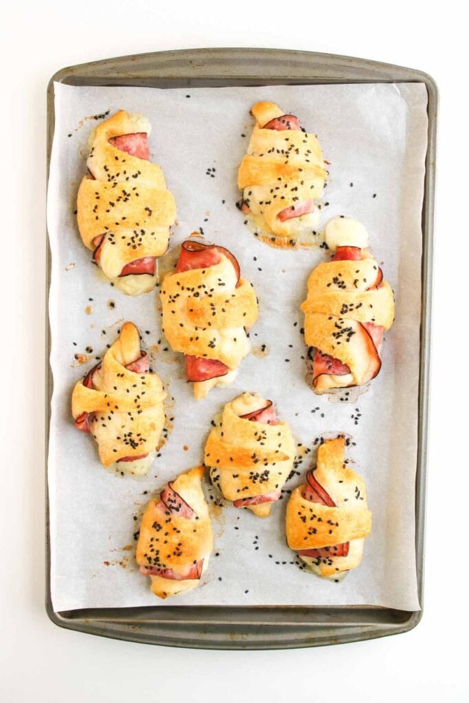 Seven Ham and Cheese Crescent Rolls, filled with savory ham and cheese and topped with black sesame seeds, are arranged on a parchment-lined baking tray.