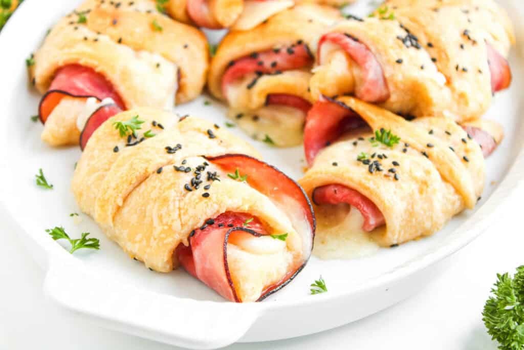Golden Ham and Cheese Crescent Rolls filled with savory ham and melted cheese, topped with black sesame seeds and parsley, beautifully arranged on a white serving dish.