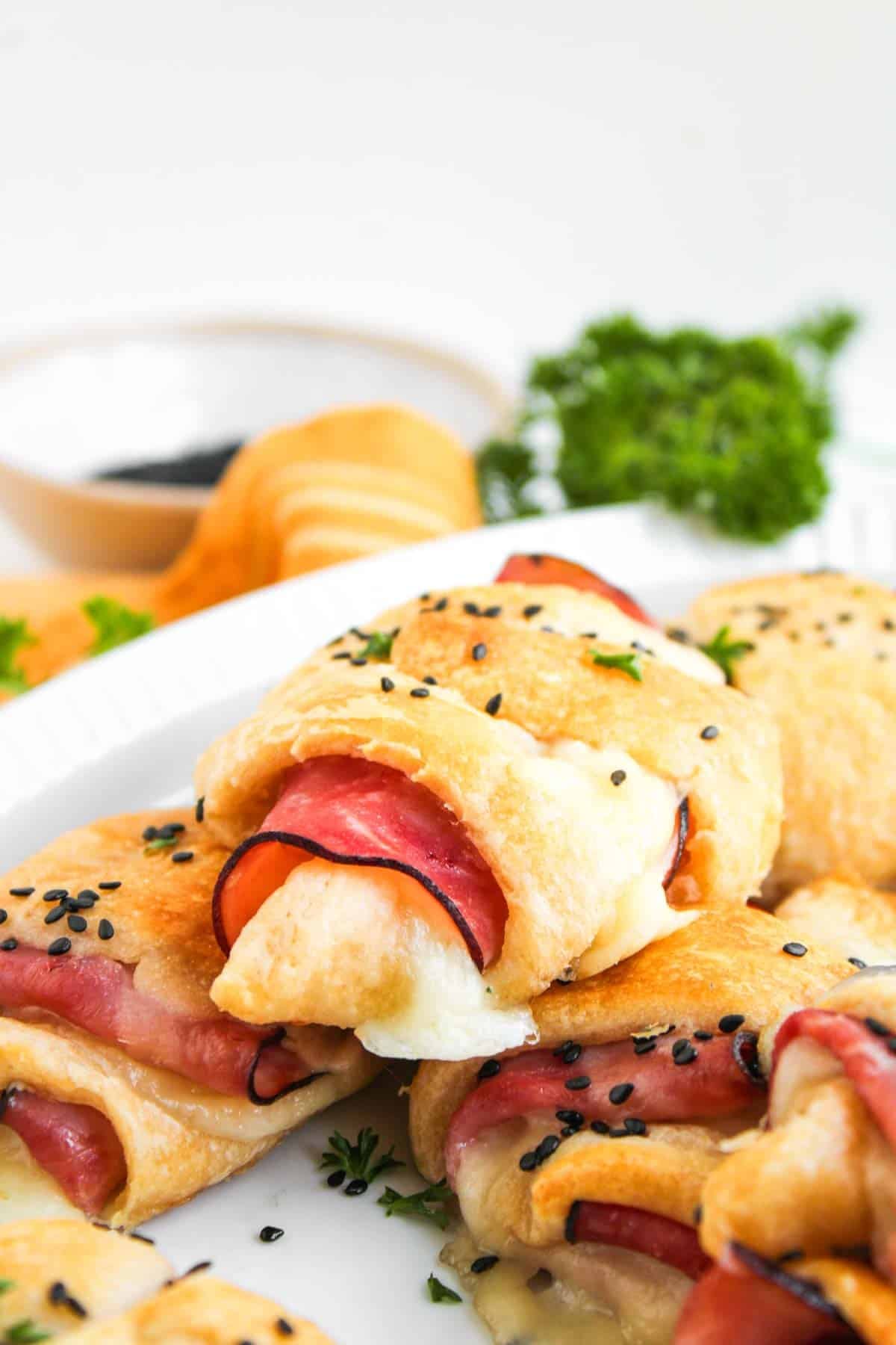 A close-up of Ham and Cheese Crescent Rolls filled with ham and melted cheese, topped with black sesame seeds, served on a white plate with parsley garnish in the background.