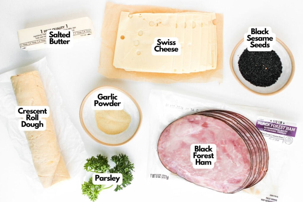 Crescent roll dough, salted butter, Swiss cheese slices, garlic powder, fresh parsley, black sesame seeds, and Black Forest ham—everything you need to make delicious Ham and Cheese Crescent Rolls—arranged on a white background.