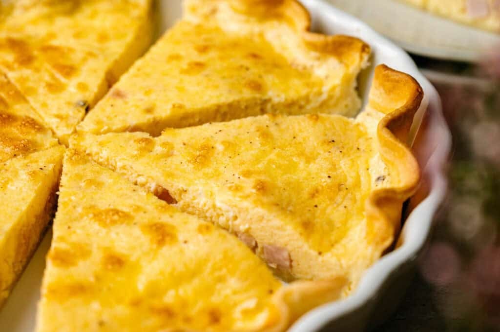 Close-up of several slices of Ham and Cheese Quiche with a golden, slightly browned top and flaky crust in a white ceramic pie dish.