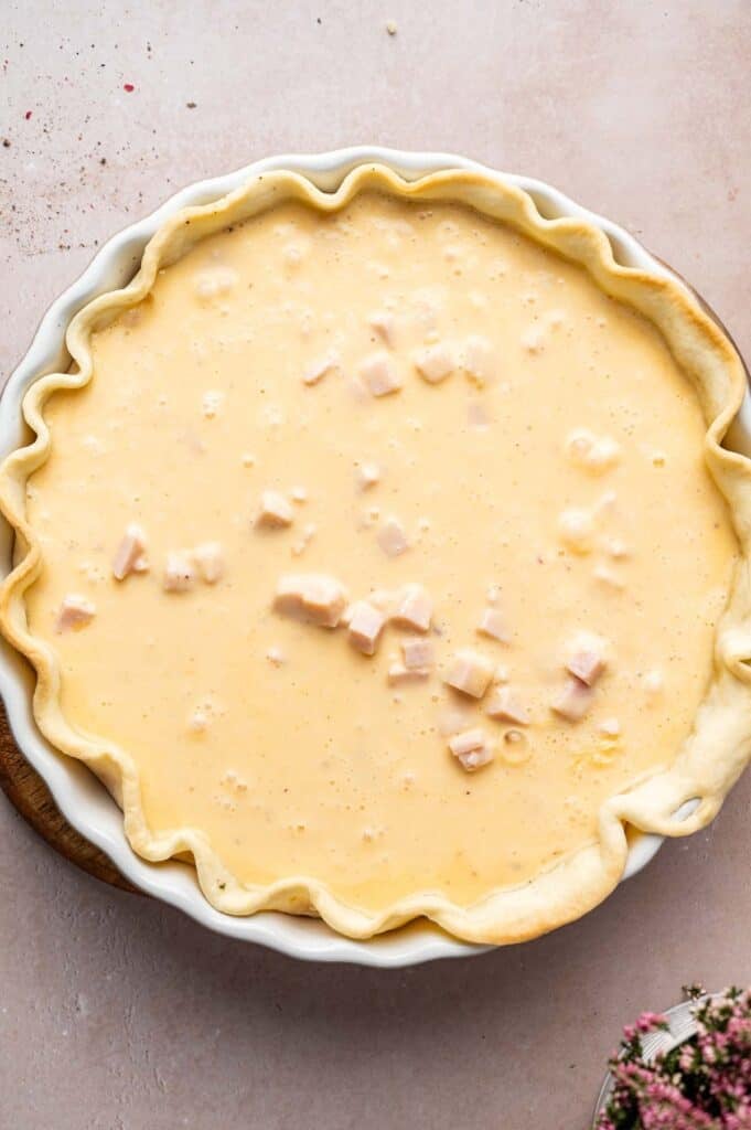 Unbaked Ham and Cheese Quiche in a pie dish with a fluted crust, filled with a creamy egg mixture and cubes of ham.