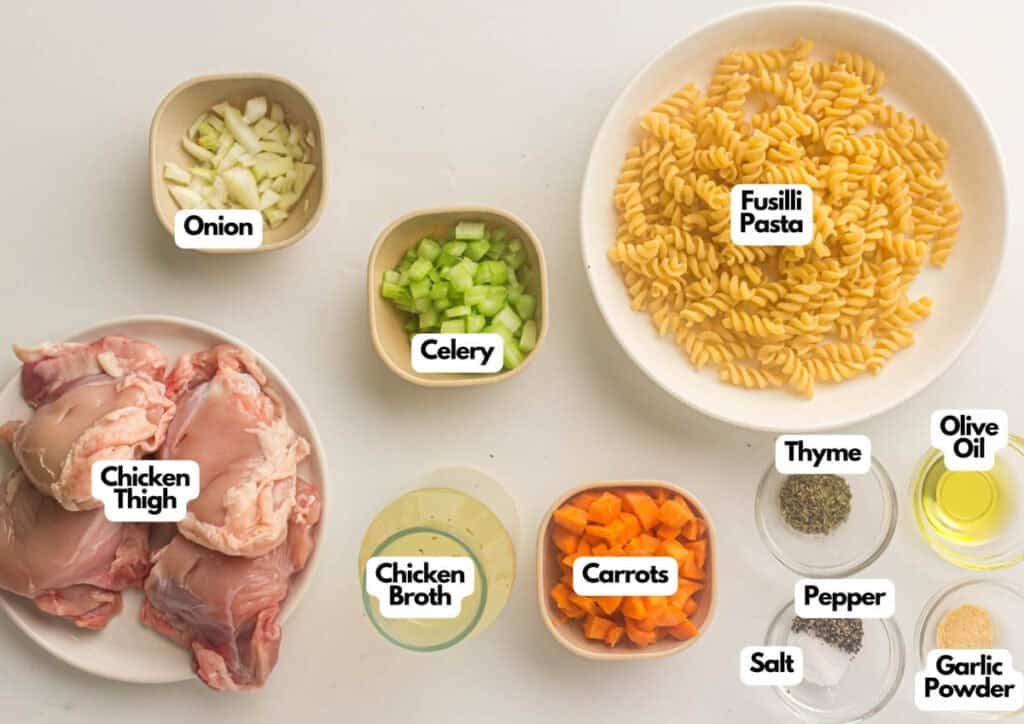 Ingredients for Instant Pot Chicken Noodle Soup arranged on a white surface, featuring chicken thighs, onion, celery, carrots, fusilli pasta, broth, olive oil, and a variety of seasonings.