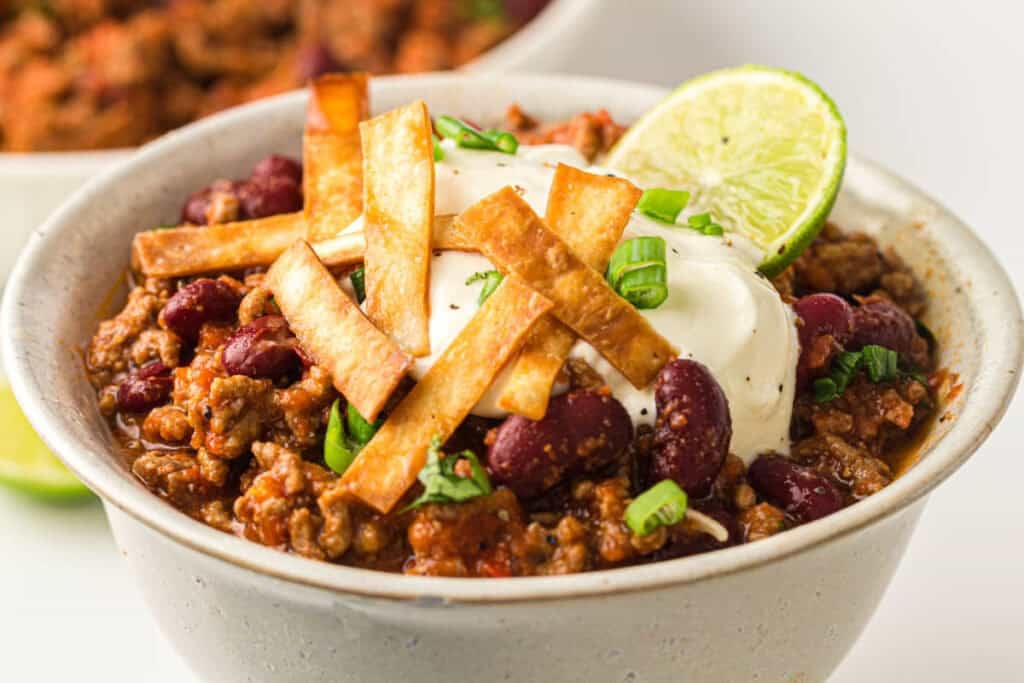 A bowl of Instant Pot Chili with kidney beans and ground meat, topped with sour cream, crispy tortilla strips, chopped green onions, and a lime slice.