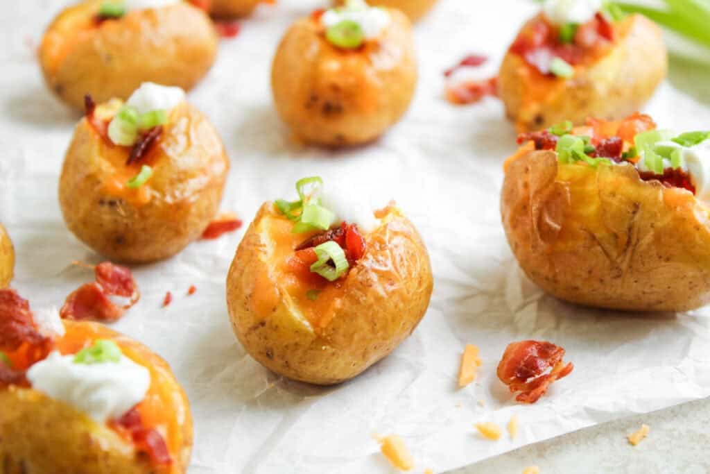 Loaded Baked Potato Bites: Mini baked potatoes topped with melted cheese, bacon bits, sour cream, and chopped green onions on parchment paper.