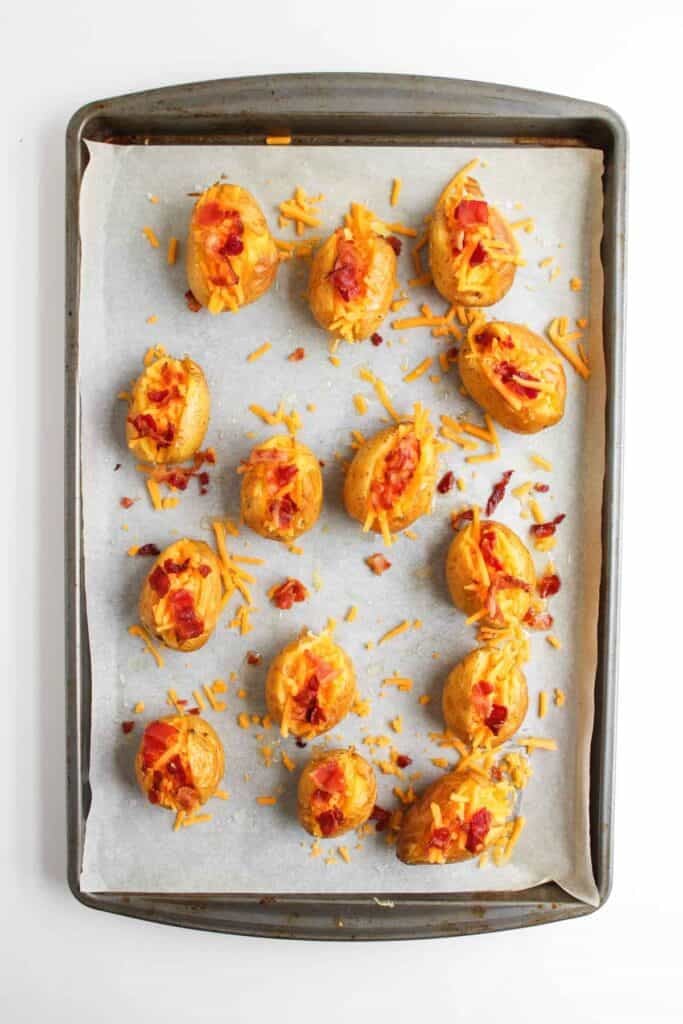 A baking sheet lined with parchment paper holds several Loaded Baked Potato Bites, each mini potato topped with shredded cheddar cheese and bacon bits.