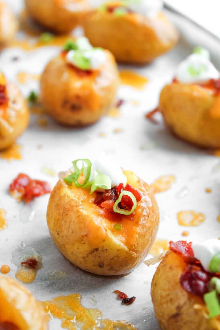 Loaded Baked Potato Bites feature mini baked potatoes topped with melted cheese, bacon bits, sour cream, and chopped green onions on a parchment-lined baking sheet.