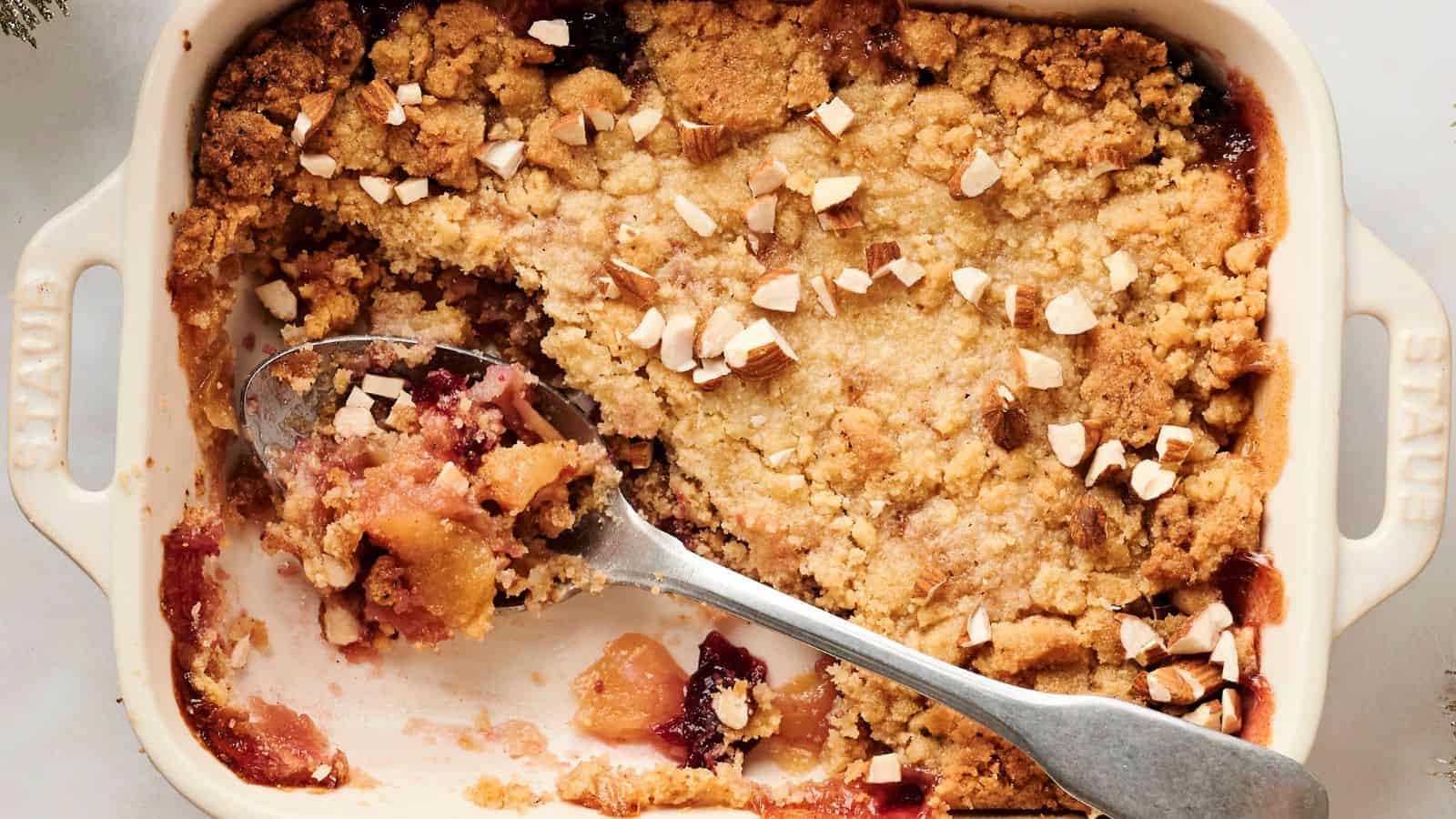 A baking dish with partially served fruit crumble topped with chopped nuts, with a serving spoon resting inside.