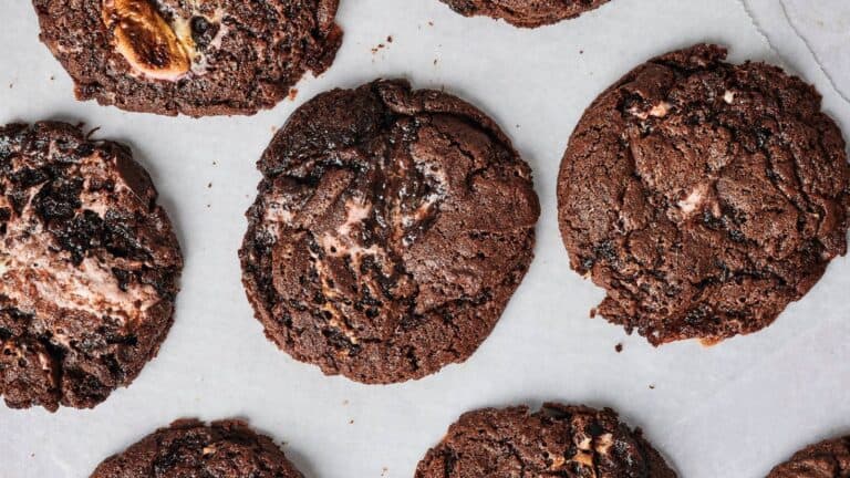 A close-up of several chocolate cookies with visible chunks and swirls, arranged on a sheet of parchment paper.