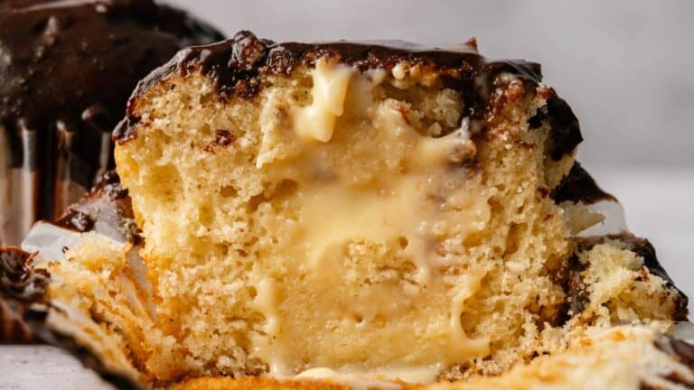 A close-up of a cross-section of a vanilla cupcake with a creamy custard filling and chocolate frosting on top.