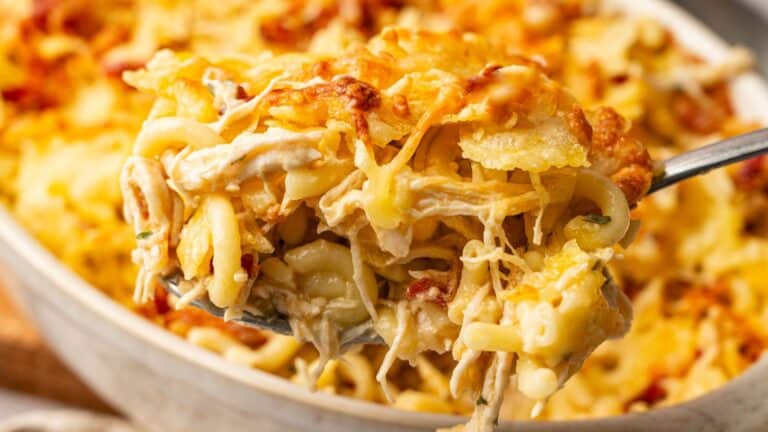 A close-up of a spoonful of cheesy baked pasta with shredded chicken and melted cheese, lifted from a casserole dish in the background.