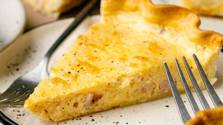 A slice of quiche with a golden crust is on a plate with a fork beside it, sprinkled with black pepper.
