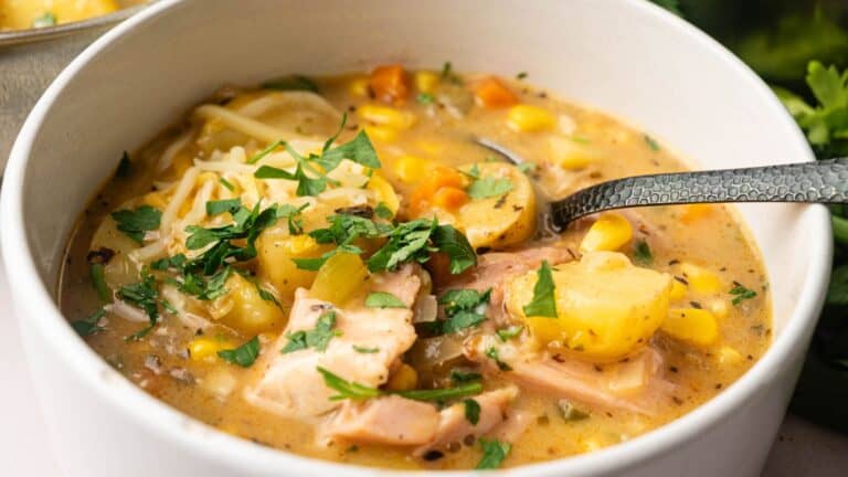 A bowl of creamy chicken and vegetable soup with potatoes, corn, carrots, and fresh parsley, with a spoon resting inside.