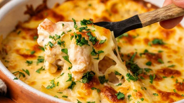 A close-up of a cheesy baked pasta dish being scooped with a spoon, topped with melted cheese and chopped fresh herbs.