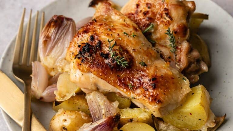 Roast chicken thighs served on a plate with golden potatoes, roasted onions, and fresh thyme, accompanied by a fork and knife.