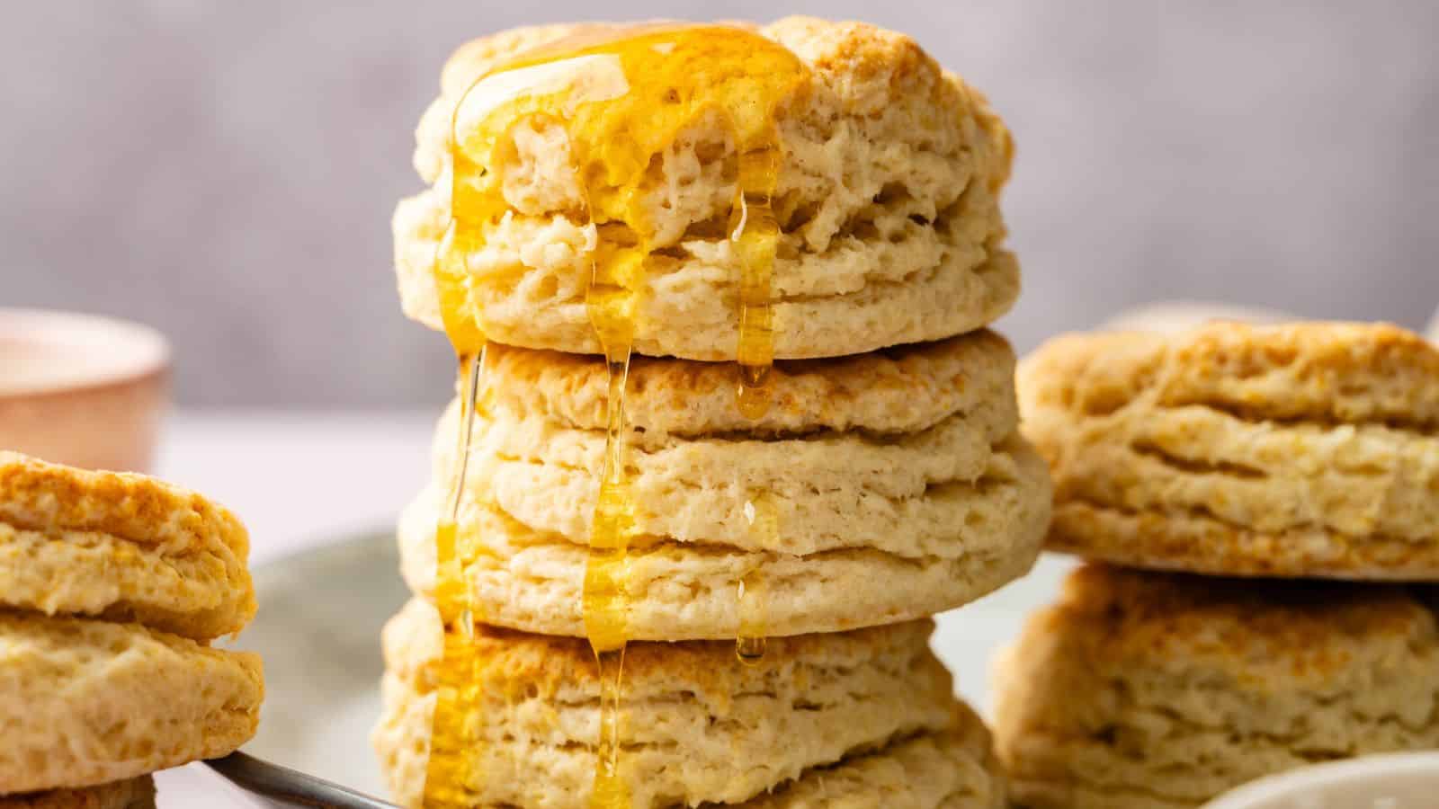 A stack of flaky biscuits with honey drizzled on top, set on a plate with more biscuits in the background.