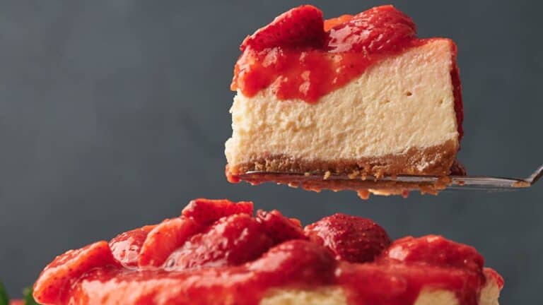 A slice of strawberry-topped cheesecake is being lifted from a whole cheesecake against a dark background.