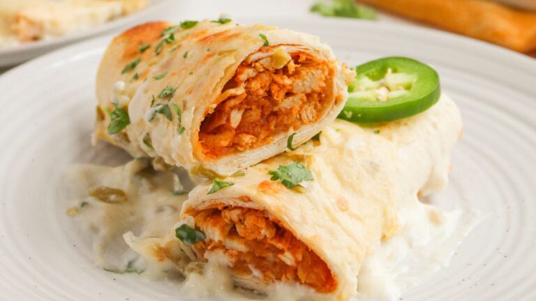 Two chicken enchiladas topped with white sauce, chopped cilantro, and a slice of jalapeño are served on a white plate.