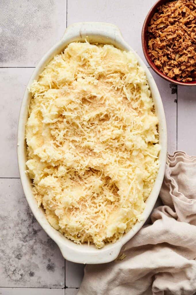 An oval baking dish filled with Mashed Potato Casserole, topped with shredded cheese and breadcrumbs, sits beside a bowl of crispy fried onions.