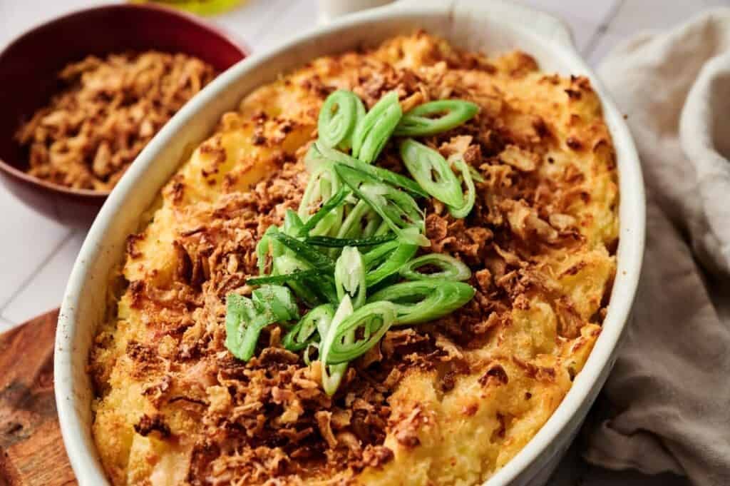 A Mashed Potato Casserole topped with crispy fried onions and sliced green onions, served in an oval dish on a table.