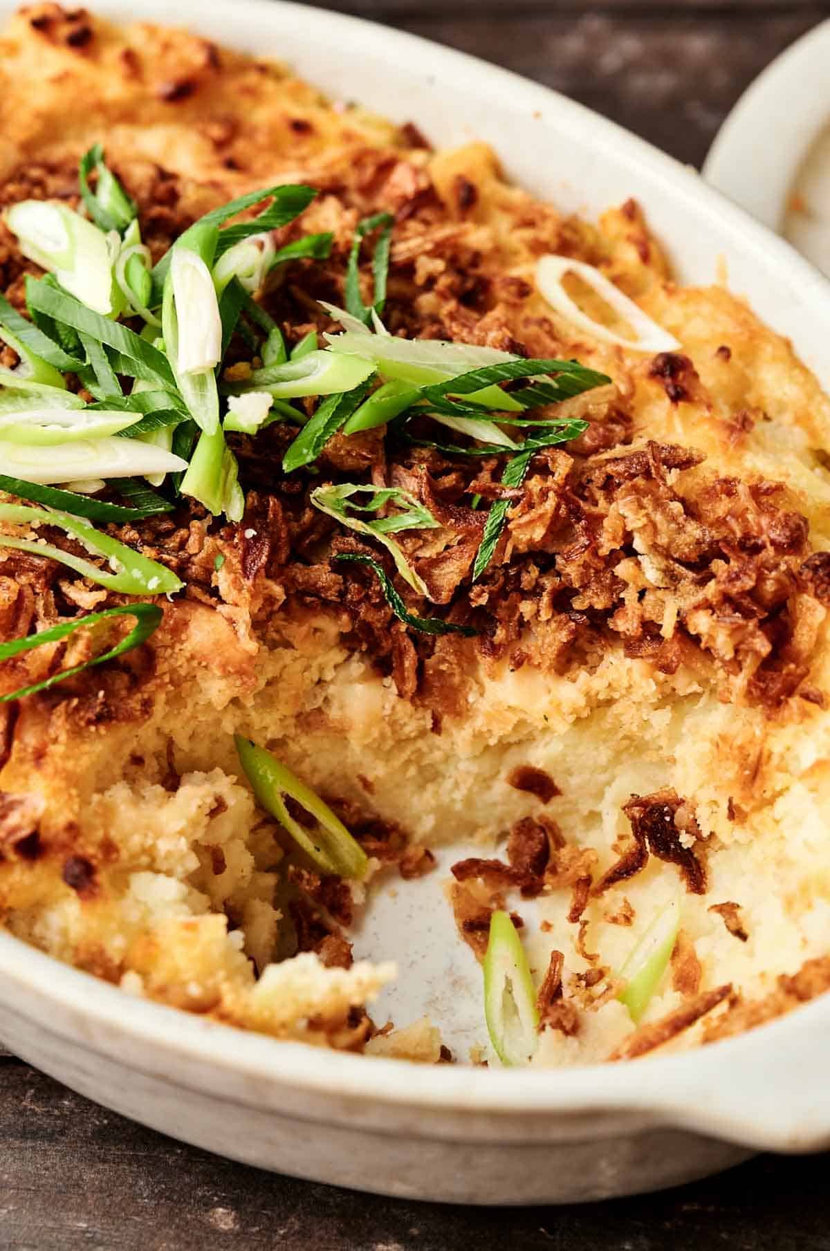 A Mashed Potato Casserole with a golden, crispy topping and sliced green onions, partially served in a white oval dish.