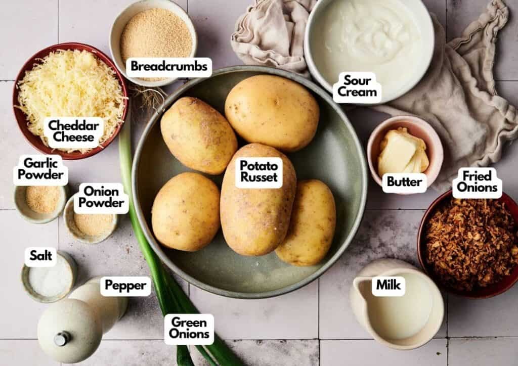 A top view of ingredients for a Mashed Potato Casserole-style twice-baked potatoes, including russet potatoes, cheddar cheese, sour cream, butter, milk, breadcrumbs, fried onions, green onions, and seasonings.