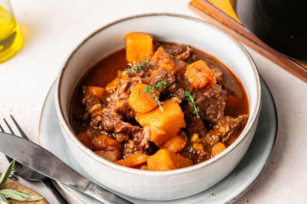 A bowl of Slow Cooker Beef Stew with tender chunks of meat, potatoes, and vegetables, garnished with fresh herbs, served on a white plate with a fork and knife beside it.