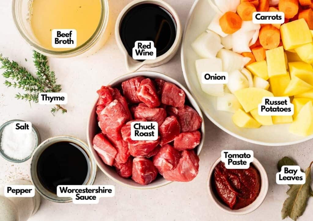 Ingredients for Slow Cooker Beef Stew are arranged on a counter, including labeled bowls of chuck roast, potatoes, carrots, onion, broth, red wine, tomato paste, spices, and sauces.
