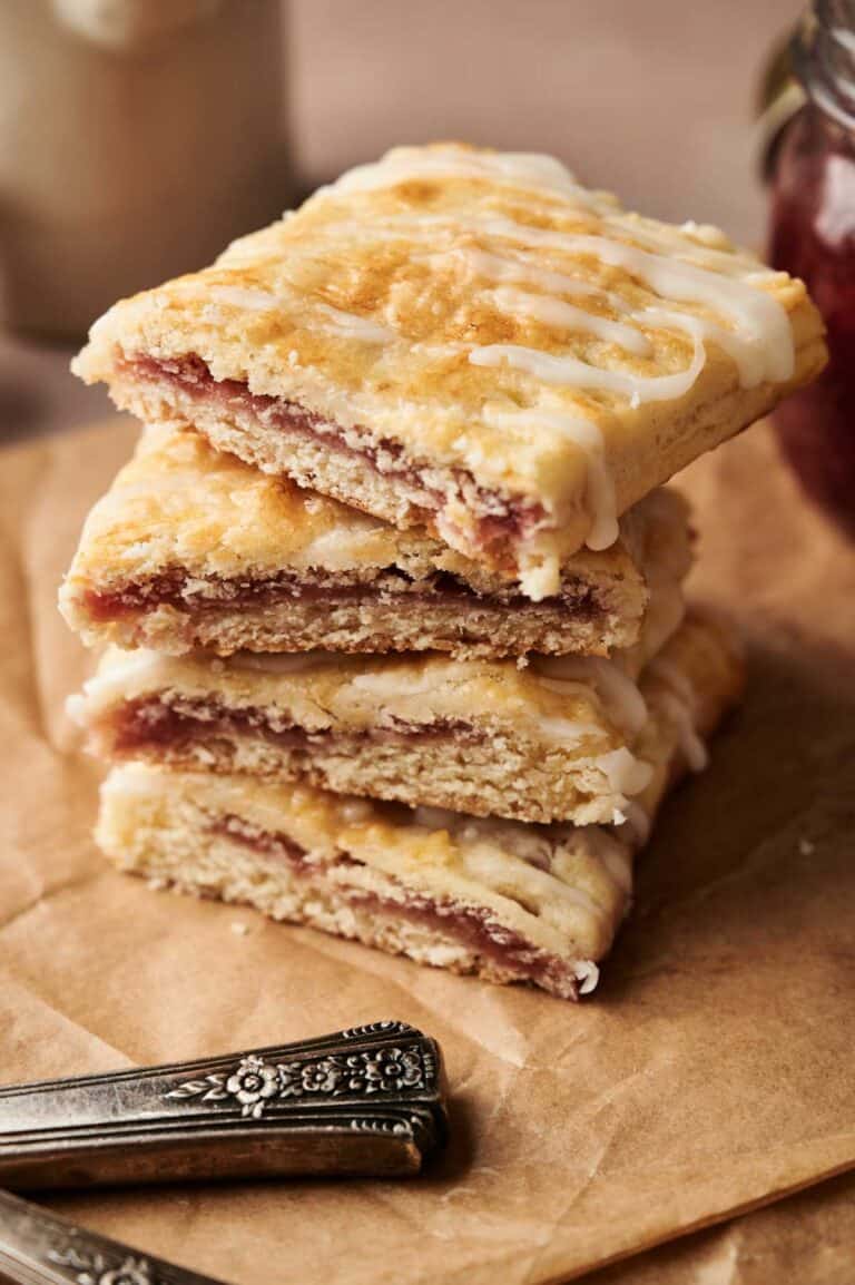 Three rectangular Sourdough Pop Tarts filled with fruit jam are stacked on brown parchment paper, with two ornate silver utensils beside them.