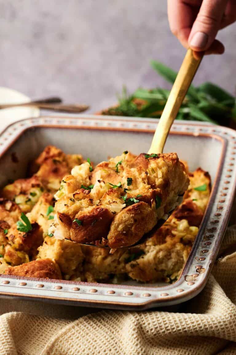 A hand uses a wooden spatula to serve a portion of baked bread stuffing from a square ceramic dish.