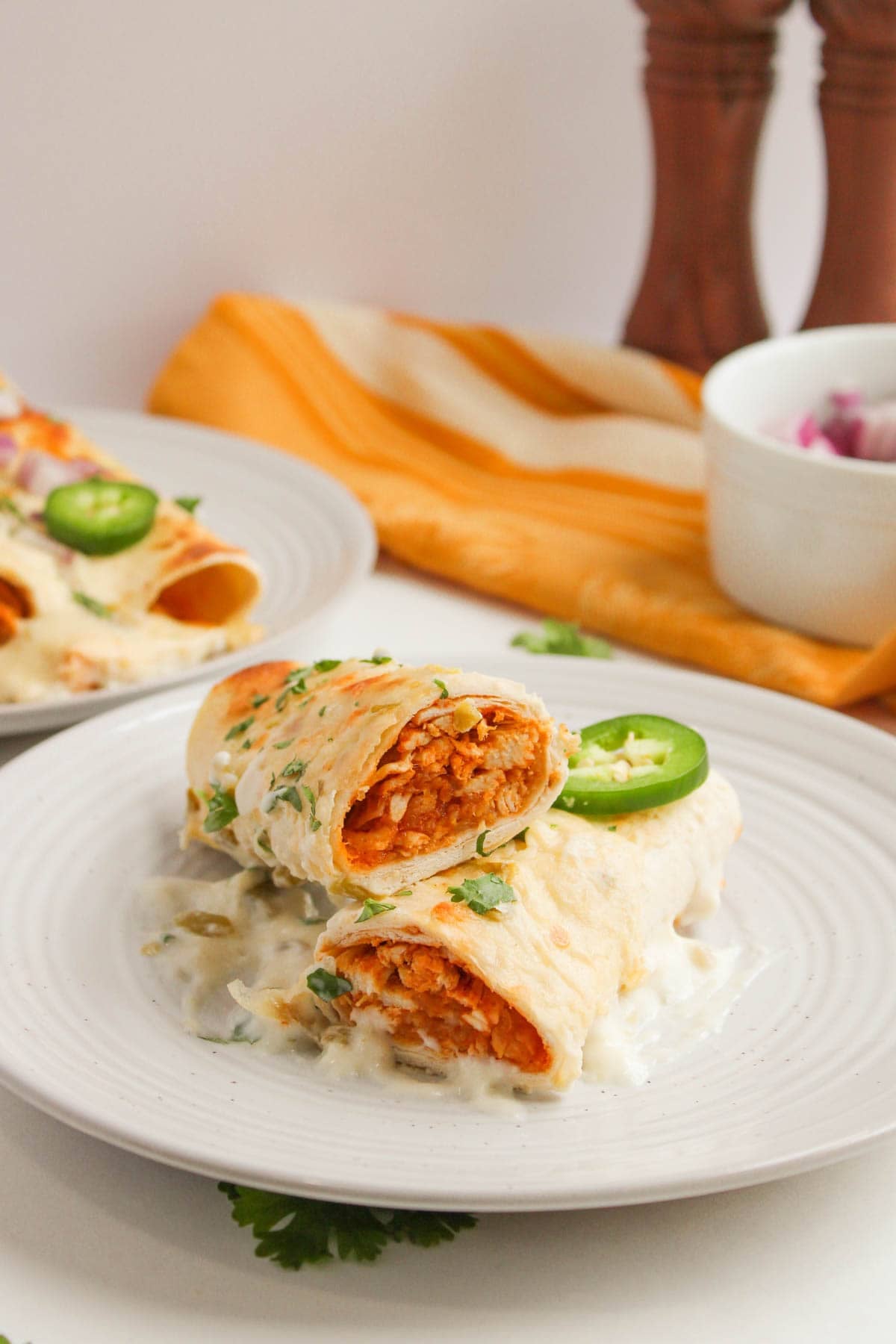 Two White Chicken Enchiladas topped with creamy white sauce, sliced jalape&ntilde;o, and fresh chopped herbs are served on a white plate, with another plate and napkin in the background.