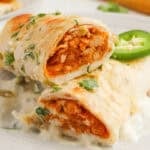 A plate with two White Chicken Enchiladas topped with creamy white sauce, garnished with chopped cilantro and a slice of jalapeño.