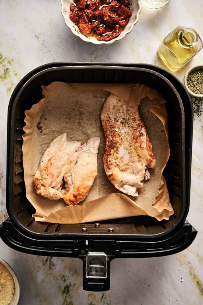 Two seasoned chicken breasts on parchment paper inside an air fryer basket, surrounded by bowls with oil, herbs, and sun-dried tomatoes&mdash;perfect for preparing Air Fryer Marry Me Chicken on a light countertop.