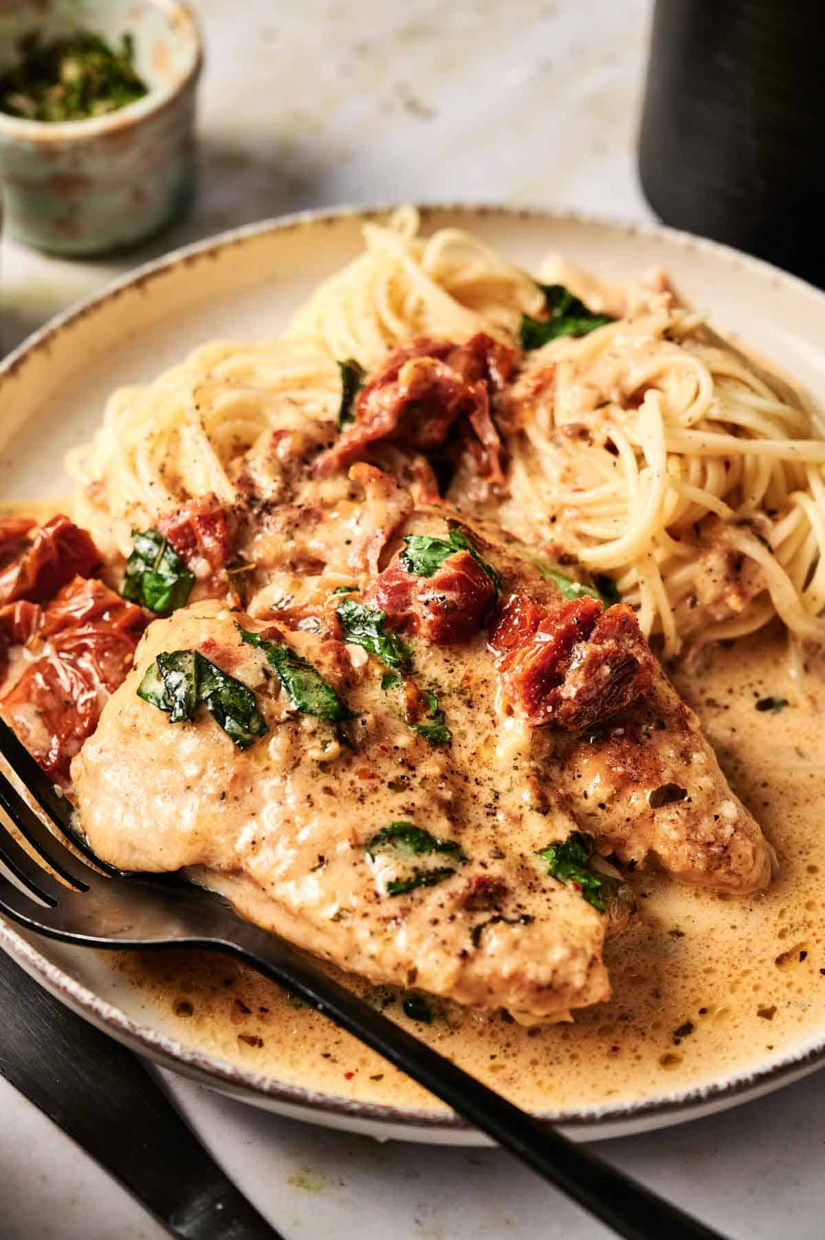 A plate of creamy Air Fryer Marry Me Chicken with sun-dried tomatoes and fresh basil, served with spaghetti, sits on a table with a fork and knife.
