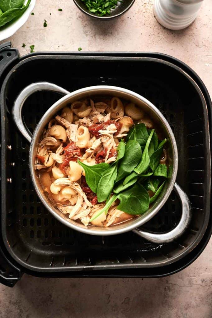 A pot of pasta, shredded chicken, tomato sauce, and fresh spinach sits inside an air fryer basket&mdash;perfect for making Air Fryer Marry Me Chicken Soup right on your countertop.
