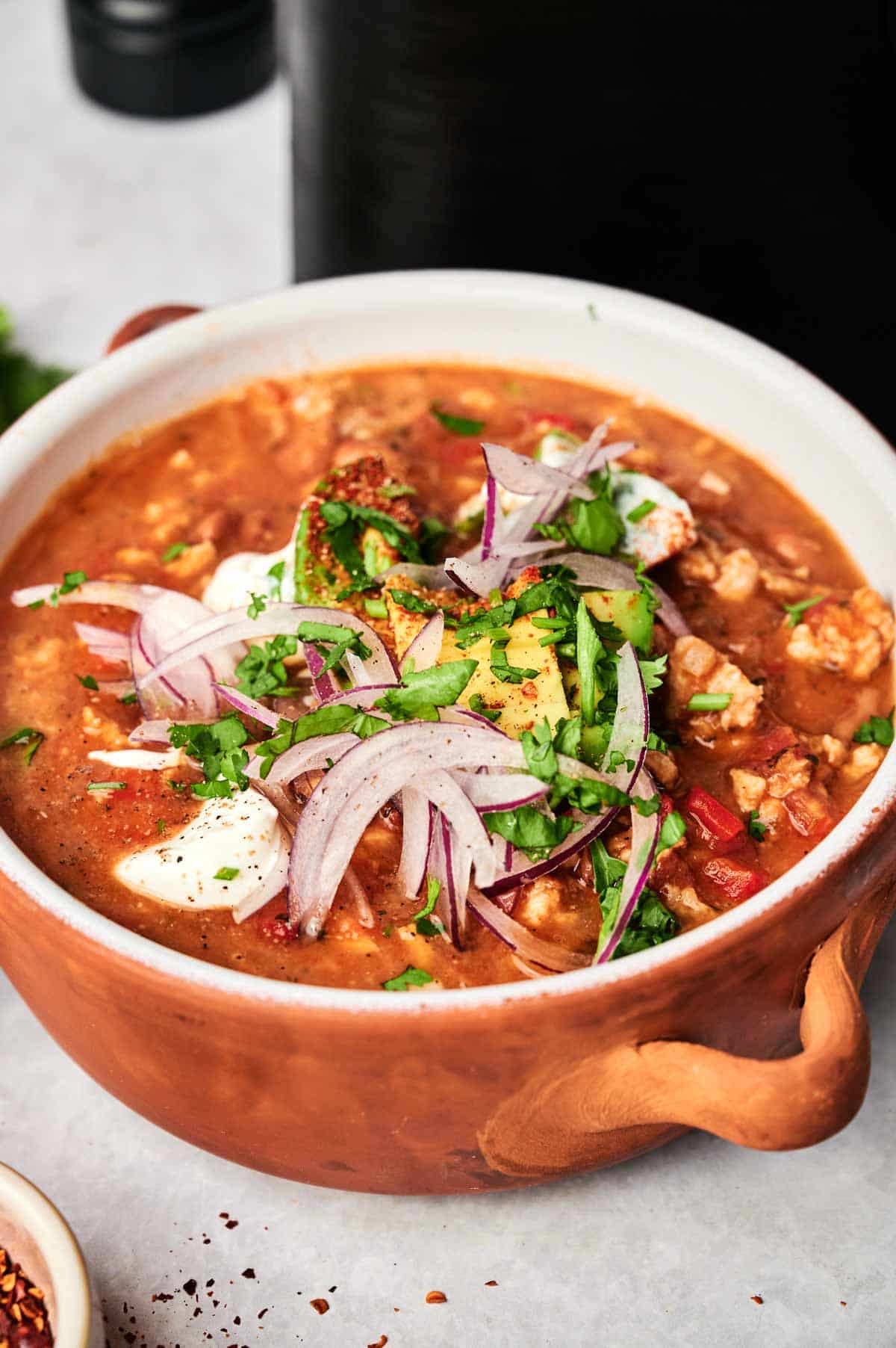A bowl of Air Fryer Turkey Chili topped with sour cream, chopped cilantro, and sliced red onions, served in a brown ceramic bowl on a light surface.