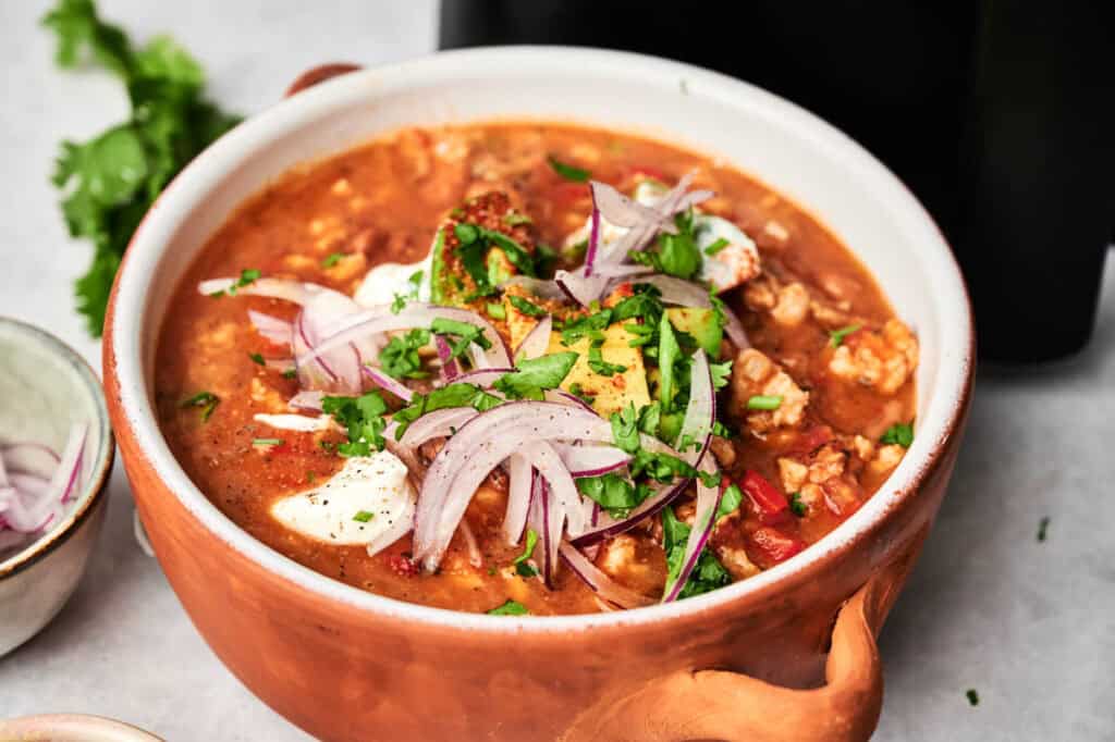 A bowl of Air Fryer Turkey Chili topped with sour cream, sliced red onions, avocado, chopped cilantro, and spices.