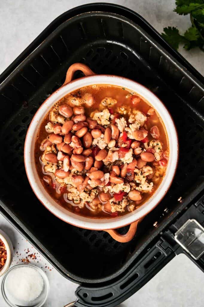 A round dish of Air Fryer Turkey Chili, filled with beans, ground turkey, and savory broth, sits inside an air fryer basket on a light-colored surface.