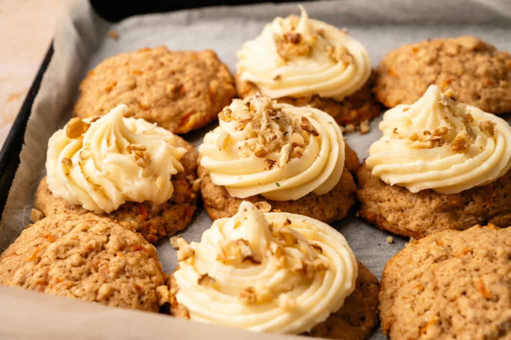 Carrot Cake Cookies topped with swirls of cream cheese frosting and chopped walnuts rest on a parchment-lined baking tray.