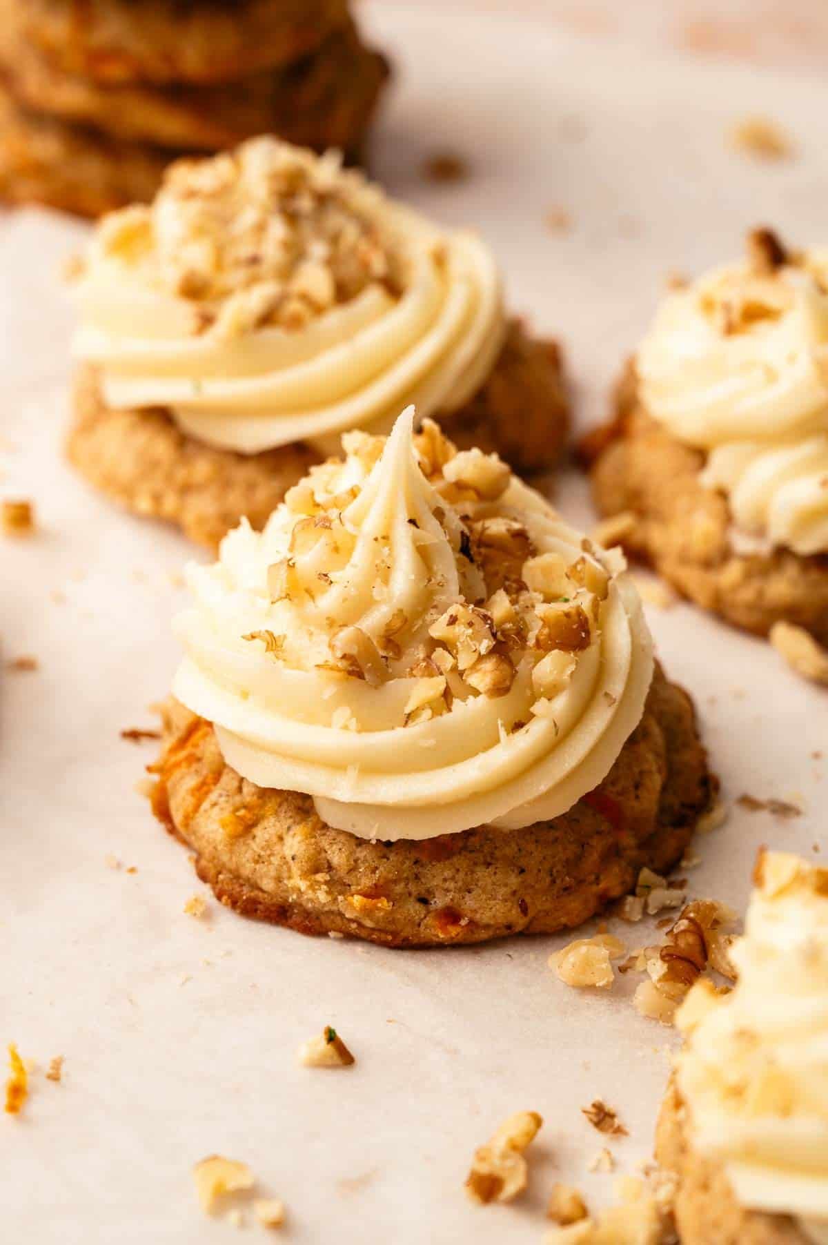 A close-up of Carrot Cake Cookies topped with swirls of frosting and sprinkled with chopped nuts on a parchment-lined surface.