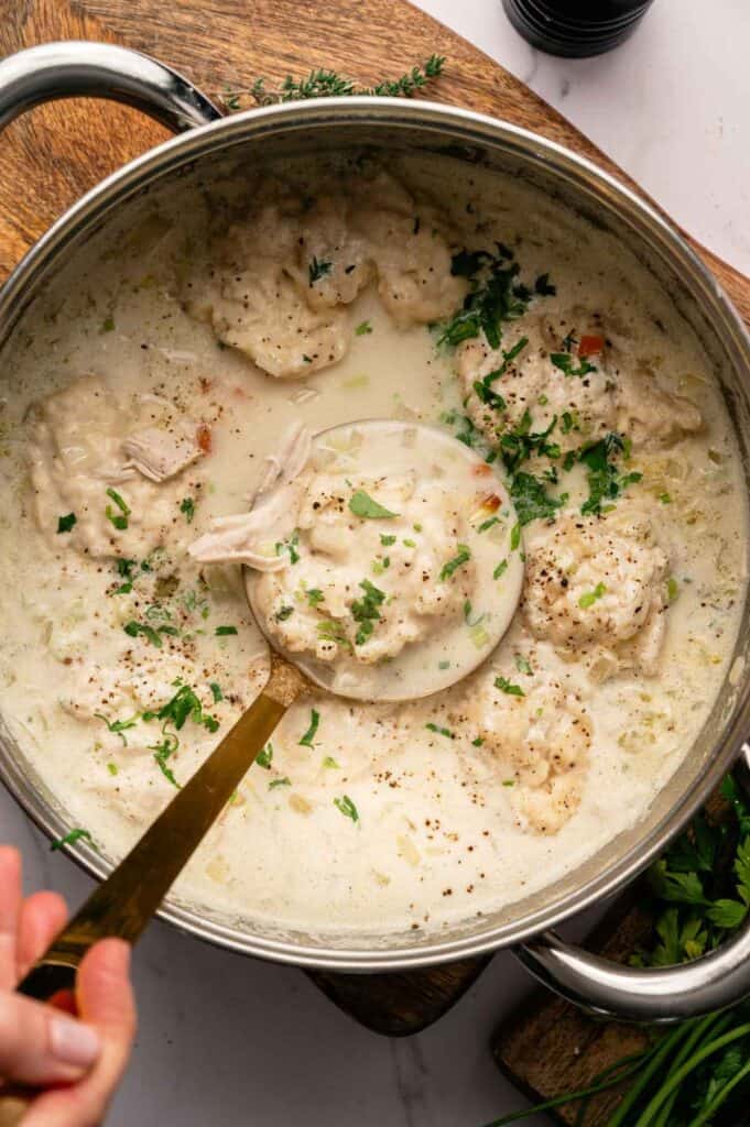 A pot of Chicken Dumpling Soup in creamy broth, garnished with chopped herbs, with a ladle lifting a hearty portion.