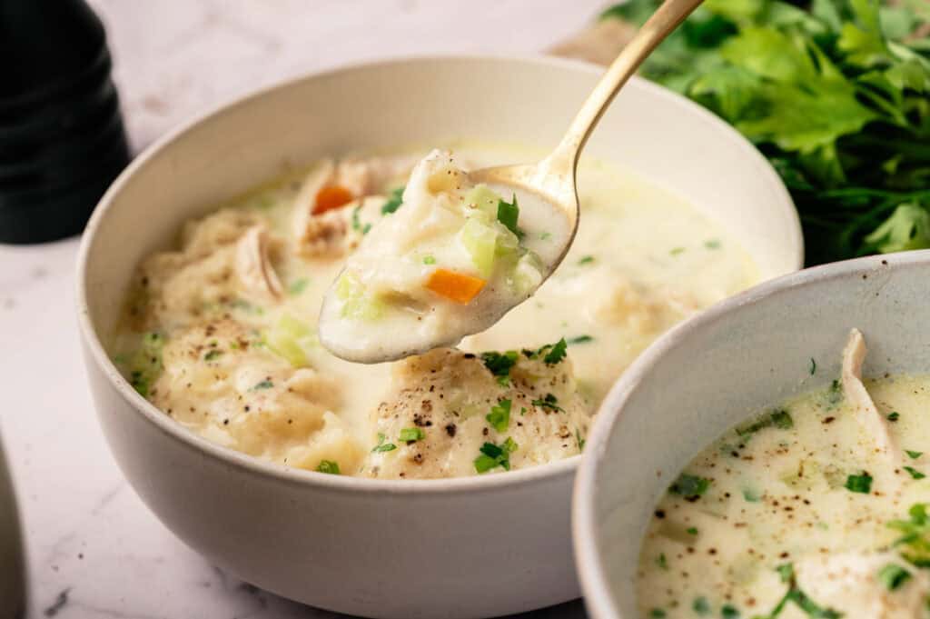 A bowl of creamy Chicken Dumpling Soup with vegetables, garnished with fresh herbs, and a spoonful being lifted from the bowl.