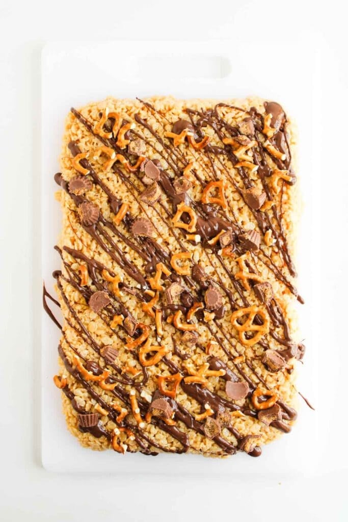 A large Chocolate Peanut Butter Rice Krispies treat topped with chocolate drizzle, mini peanut butter cups, and pretzel pieces sits on a white cutting board.