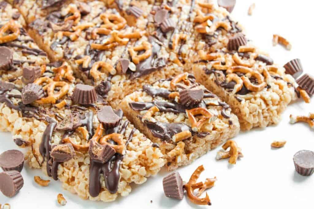 Chocolate Peanut Butter Rice Krispies treats topped with pretzel pieces, chocolate drizzle, and mini peanut butter cups, cut into squares on a white surface.