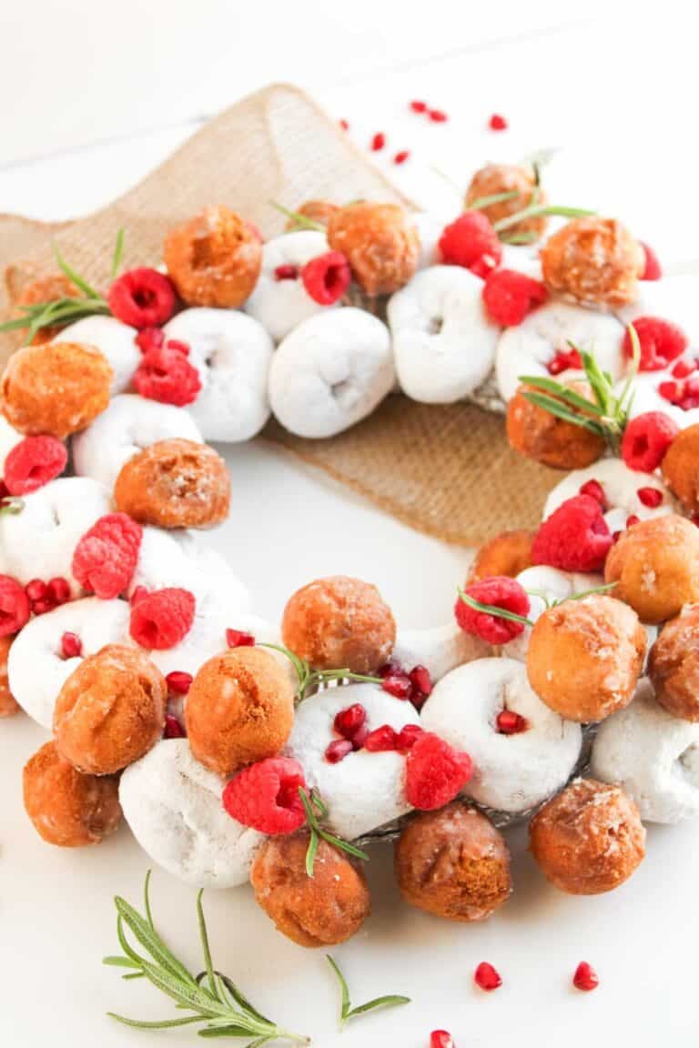 A Christmas Donut Wreath made of powdered and glazed donut holes, adorned with fresh raspberries and rosemary sprigs, is beautifully displayed on a piece of burlap.