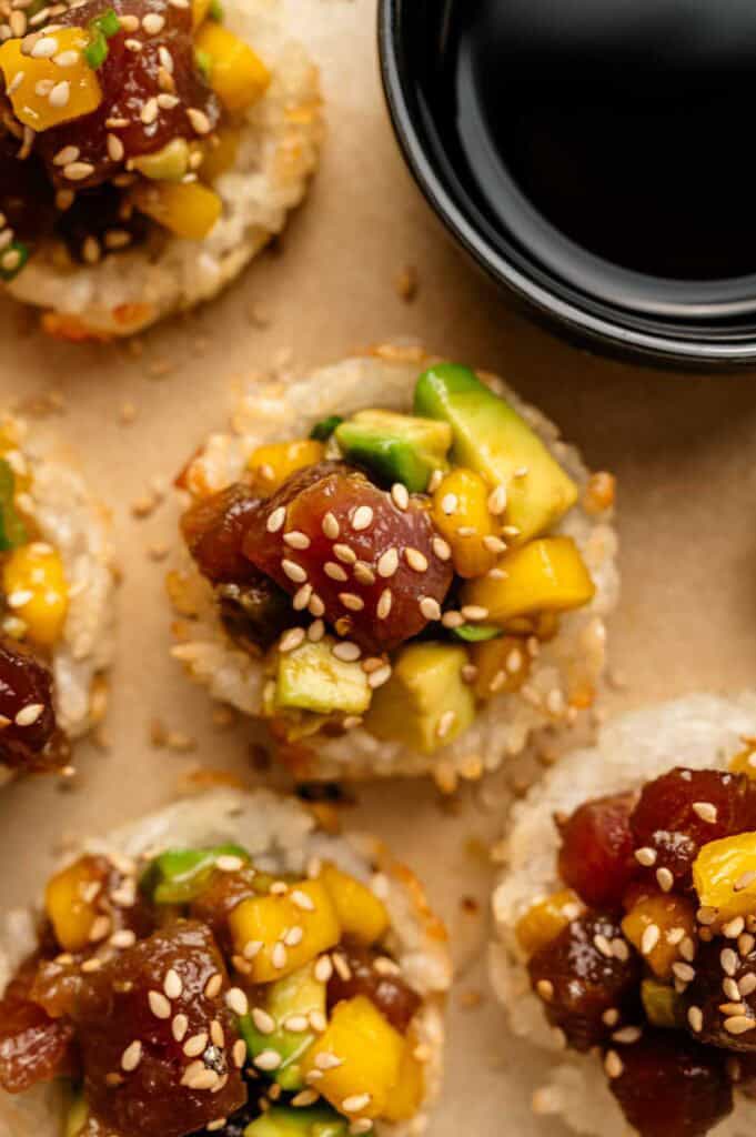 Close-up of Spicy Tuna Crispy Rice bites topped with diced avocado, mango, and sesame seeds, served beside a small bowl of soy sauce.