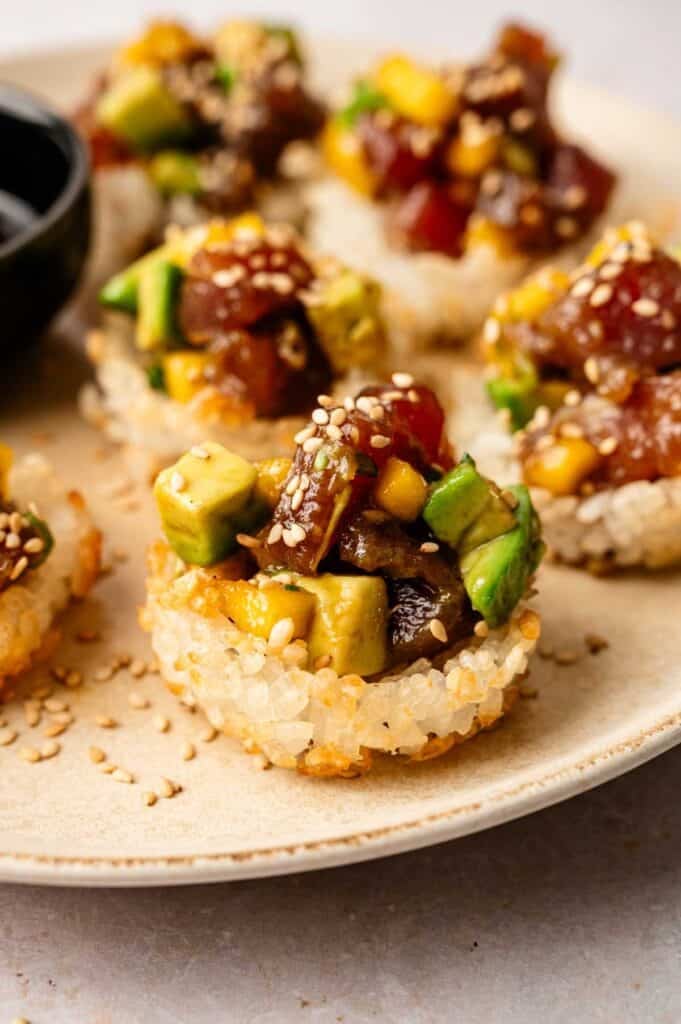 Crispy rice rounds topped with spicy tuna crispy rice, diced avocado, raw fish, mango, and sesame seeds are artfully arranged on a beige plate.
