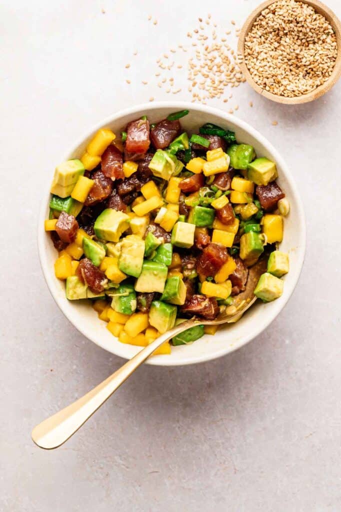 A bowl of poke salad with diced avocado, mango, tuna, and green onions, served with a spoon and a small bowl of sesame seeds&mdash;perfect alongside Spicy Tuna Crispy Rice for a vibrant meal.