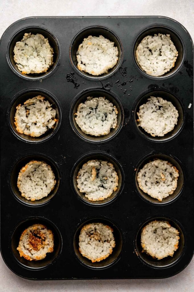A muffin tin with twelve cups, each lined with a layer of cooked rice forming shallow bowls, some slightly browned around the edges, perfect for making Spicy Tuna Crispy Rice bites.